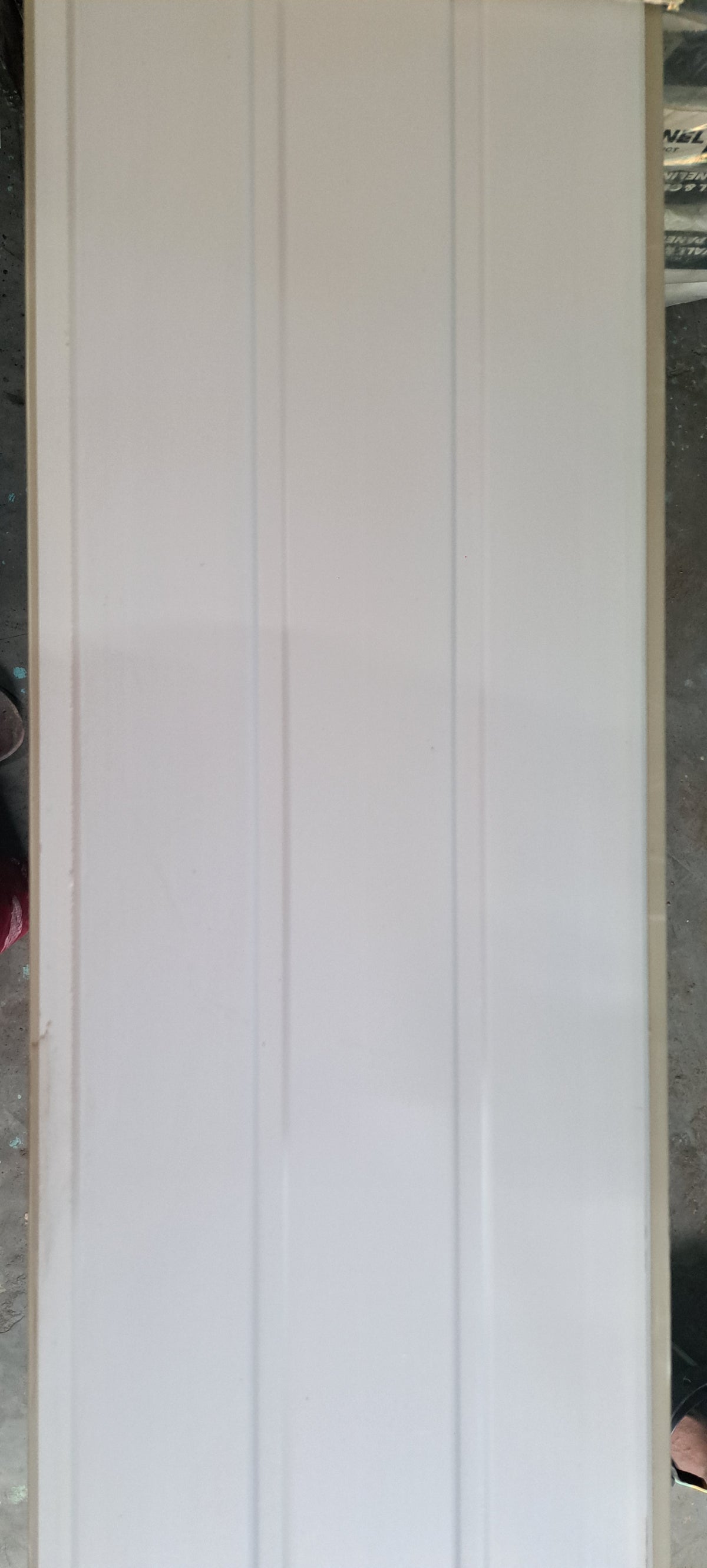 A tall, narrow white panel with three vertical grooves, photographed indoors against a gray floor and surrounding objects.